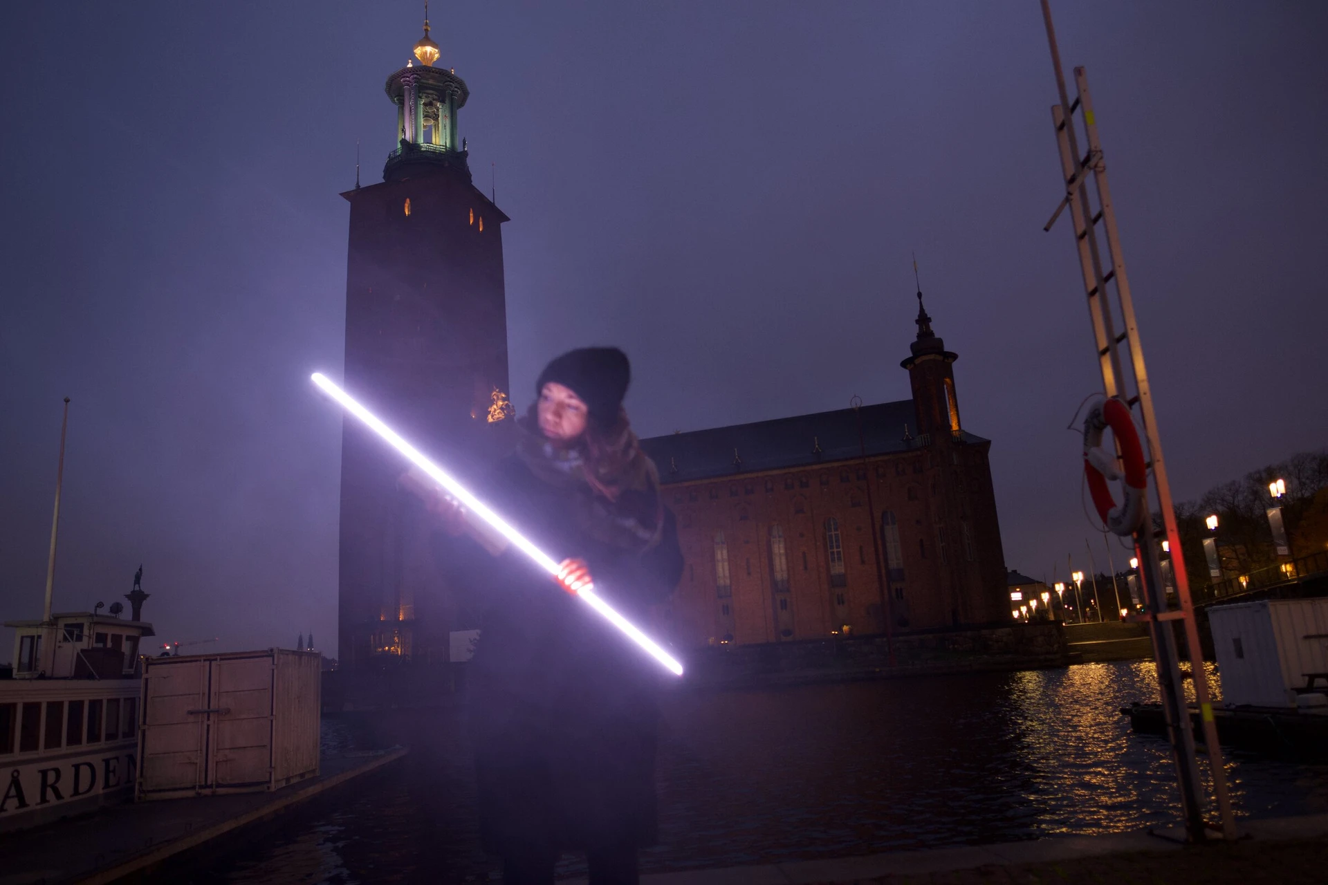 A person holds the glowing LED stick on the quay in front of Stockholm City Hall at dusk.