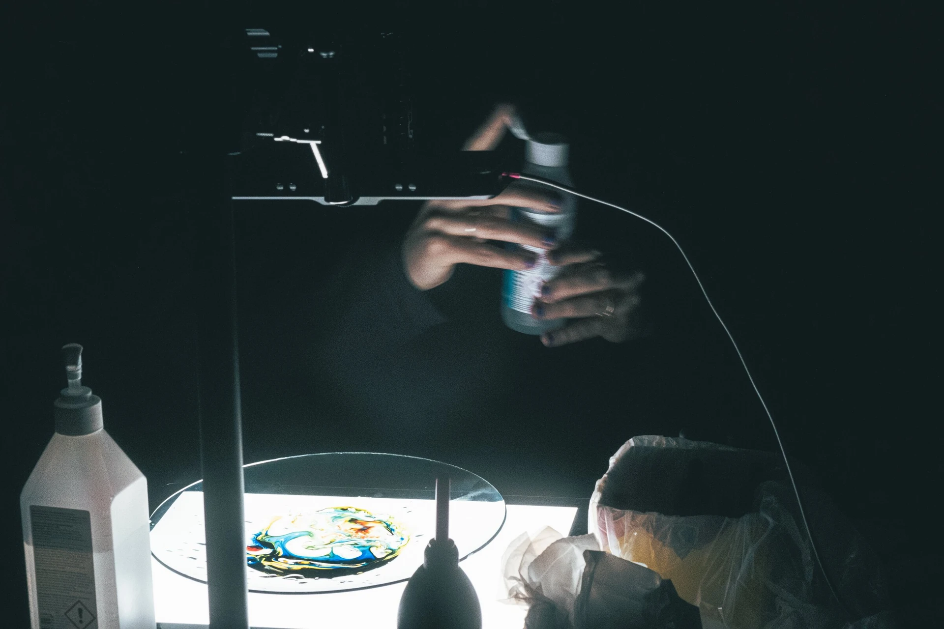 Hands pour liquid colour onto a glass dish on a light table, under a camera on a stand.
