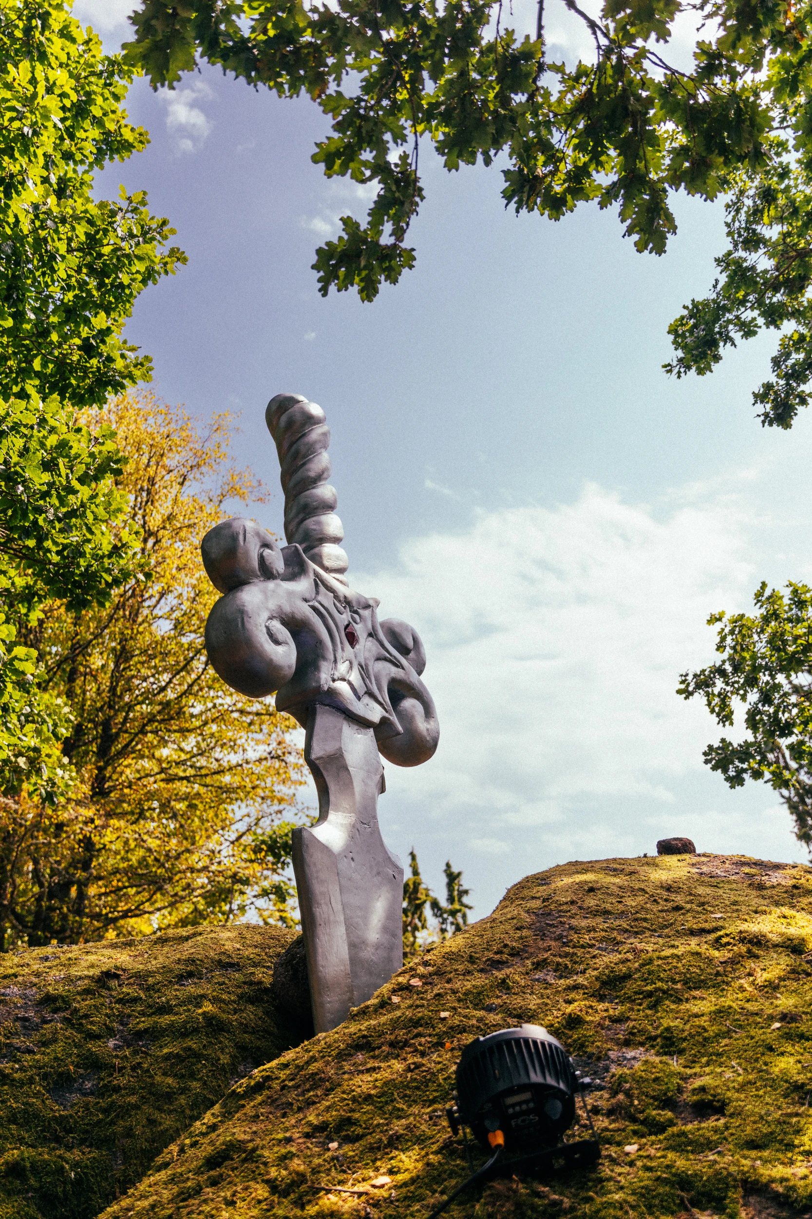 A pale sculpture of coiled forms stands on a mossy granite outcrop under an oak, with a stage light at its foot.