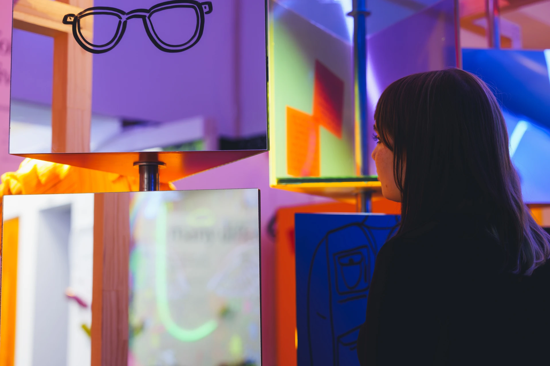 A visitor looks into reflective display boxes edged with orange and pink light.