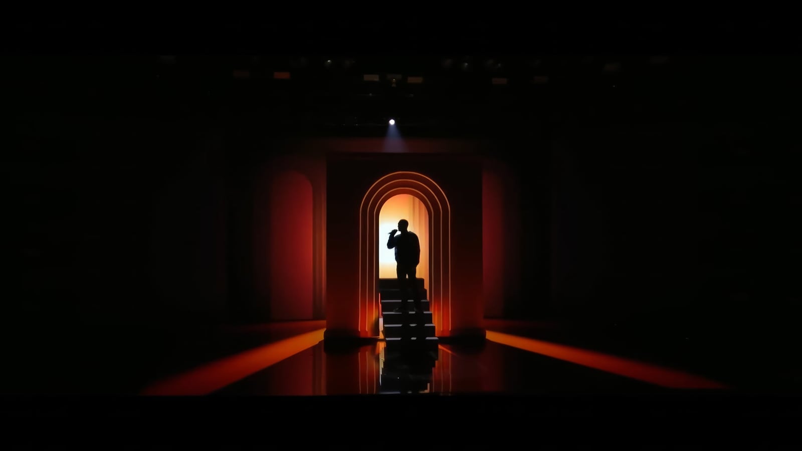 A silhouetted performer stands inside nested orange arches that extend into the dark stage.