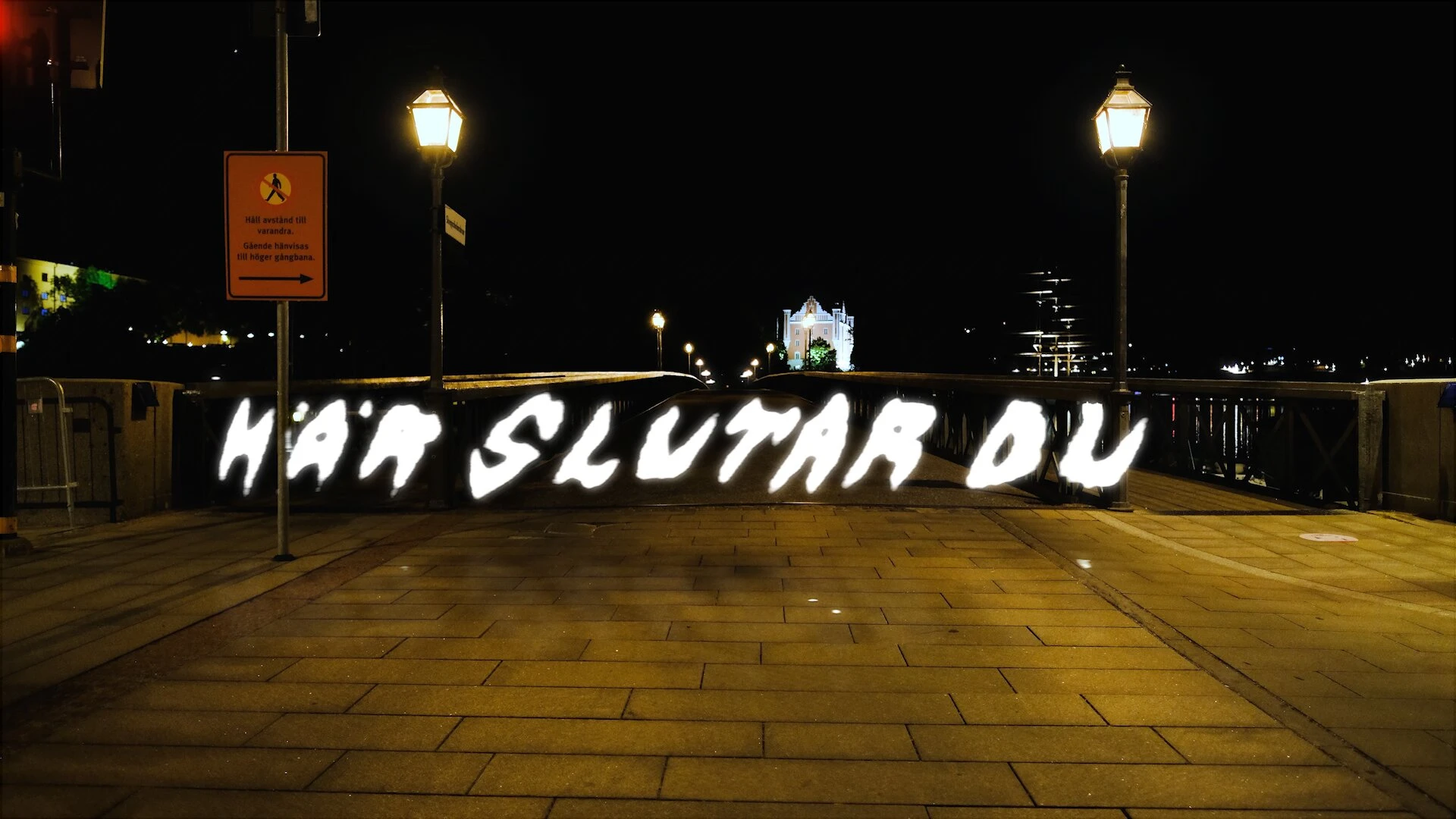Här slutar du, written in light on a walkway at night beside an orange sign asking people to keep their distance.