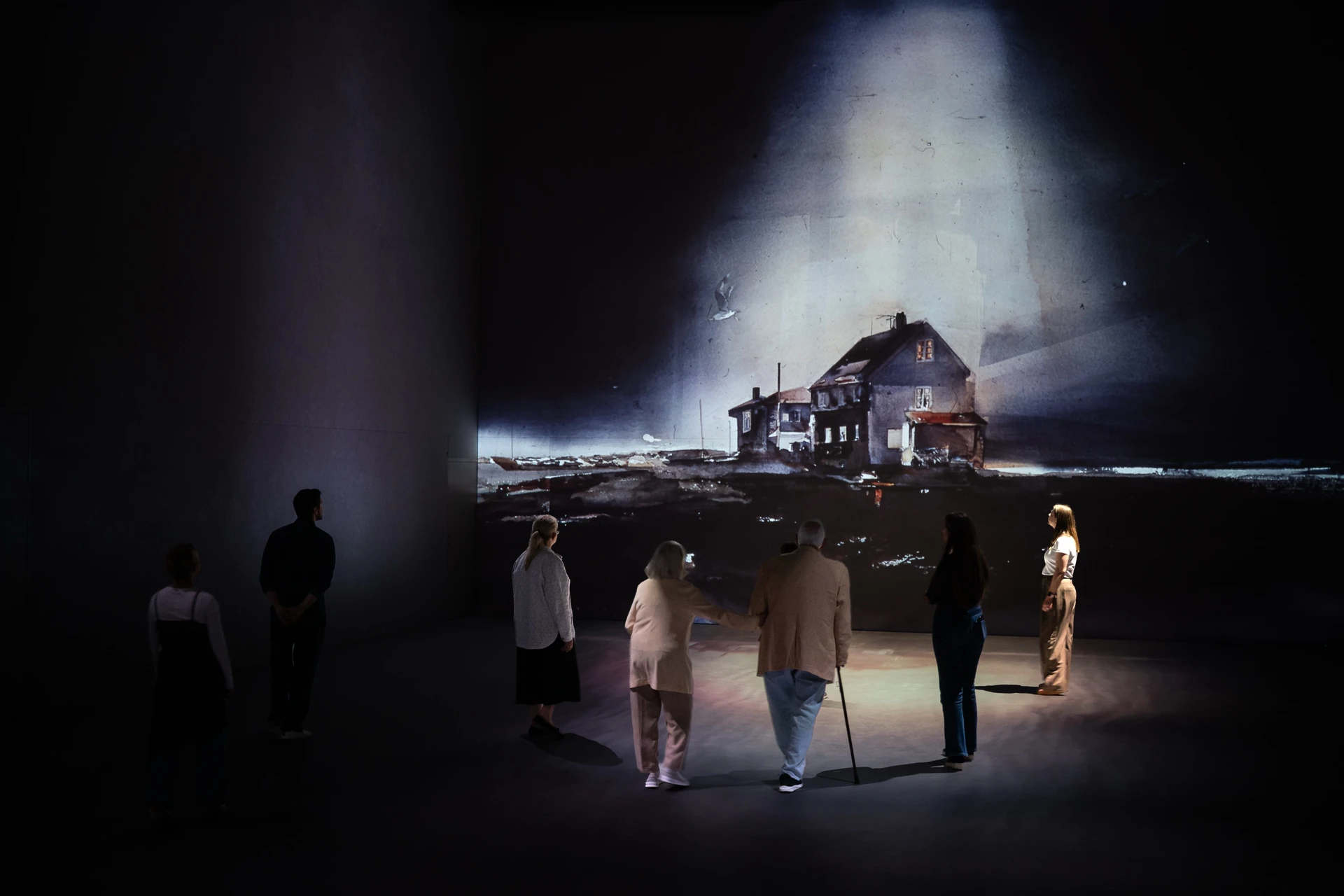 A small group stands in the dark hall facing a single projected watercolour of a house by the sea, lit from above.