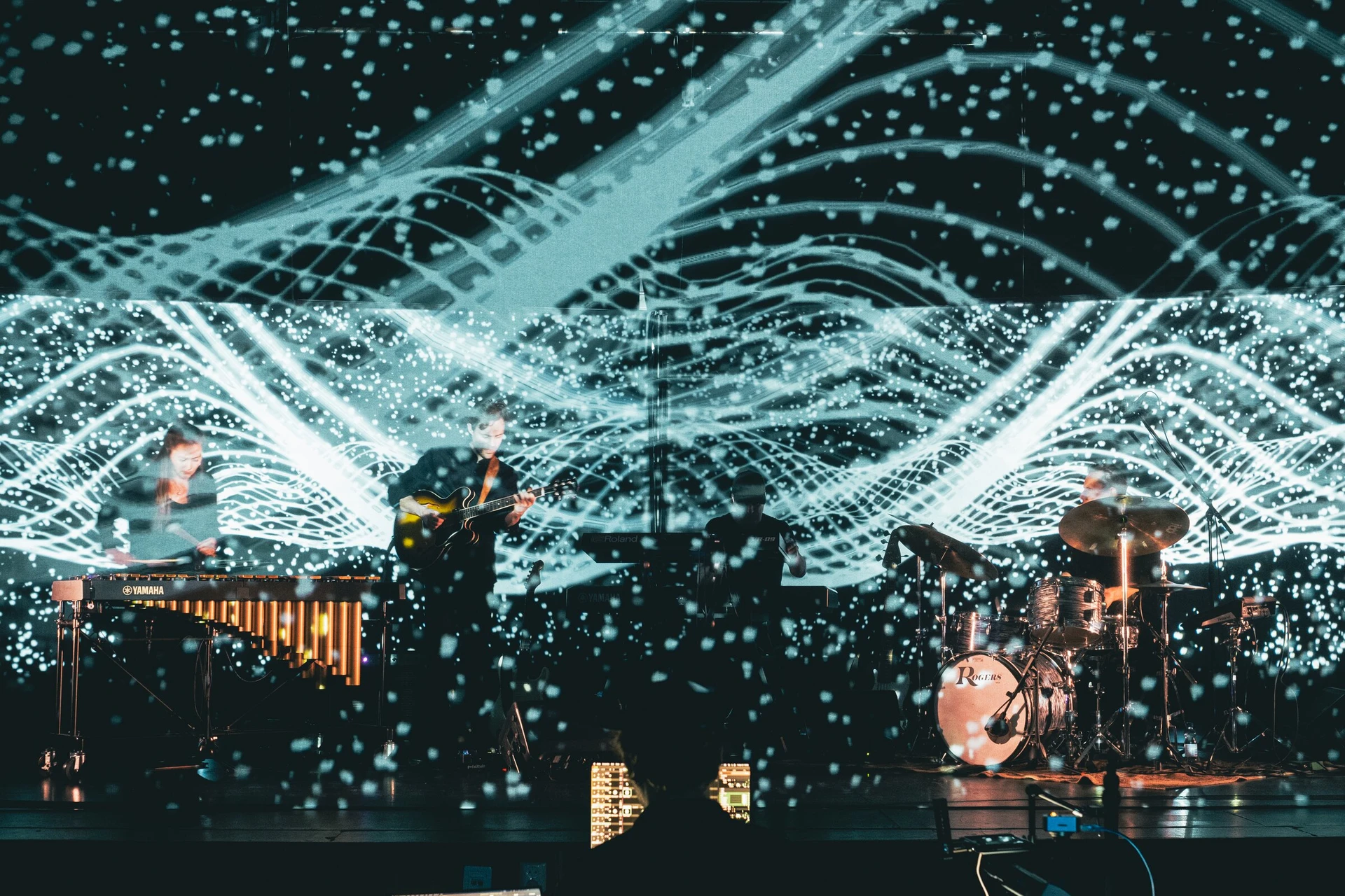 Waves of white particles fill a gauze and the screen behind it, with a vibraphonist, guitarist and drummer playing between the two layers.