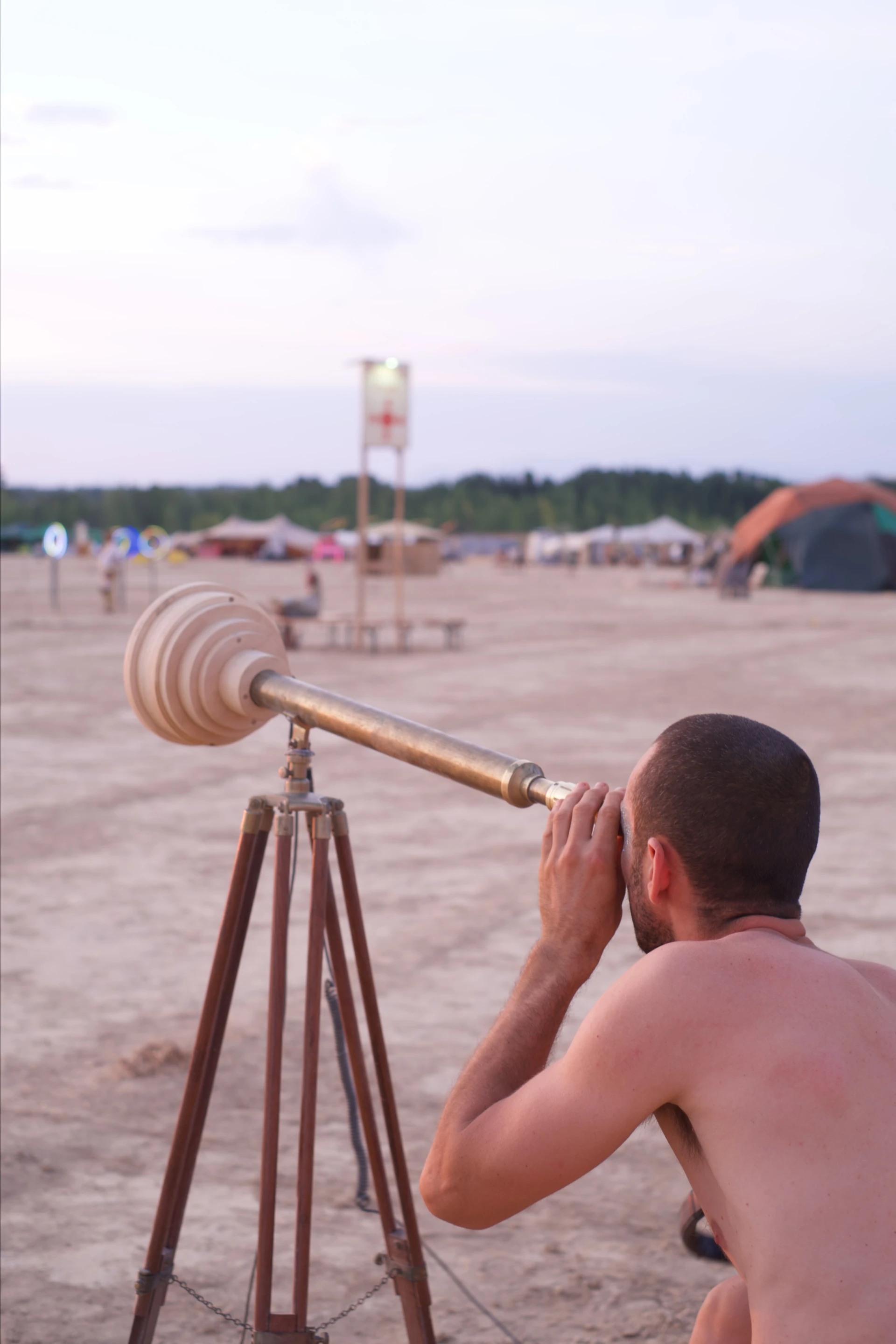 A visitor looks through the brass telescope at dusk, seen from behind with its wooden housing and tripod in view.