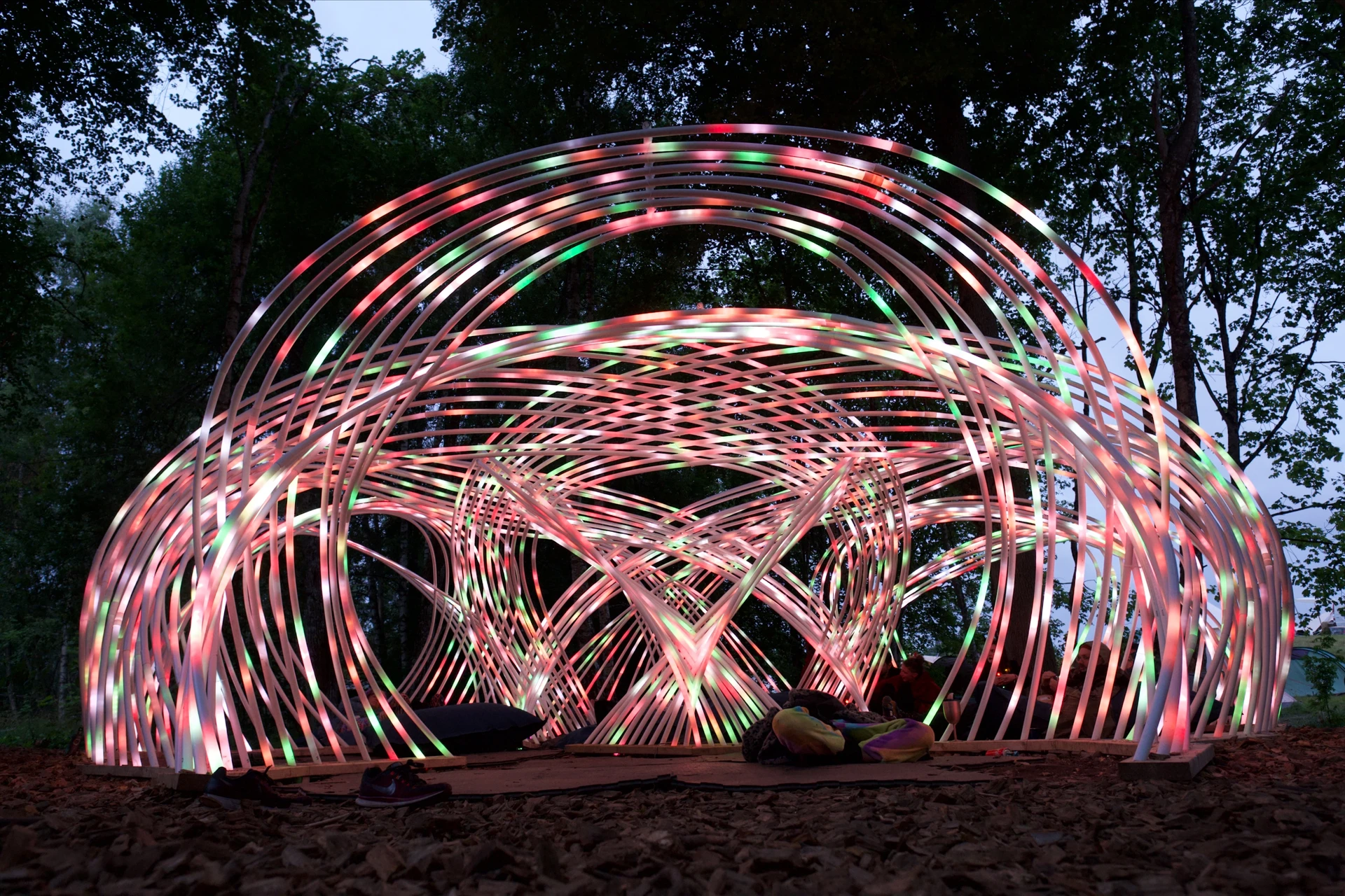 A frontal view of Kagora at dusk, with pink, green and white light tracing the woven arches above visitors resting on cushions.