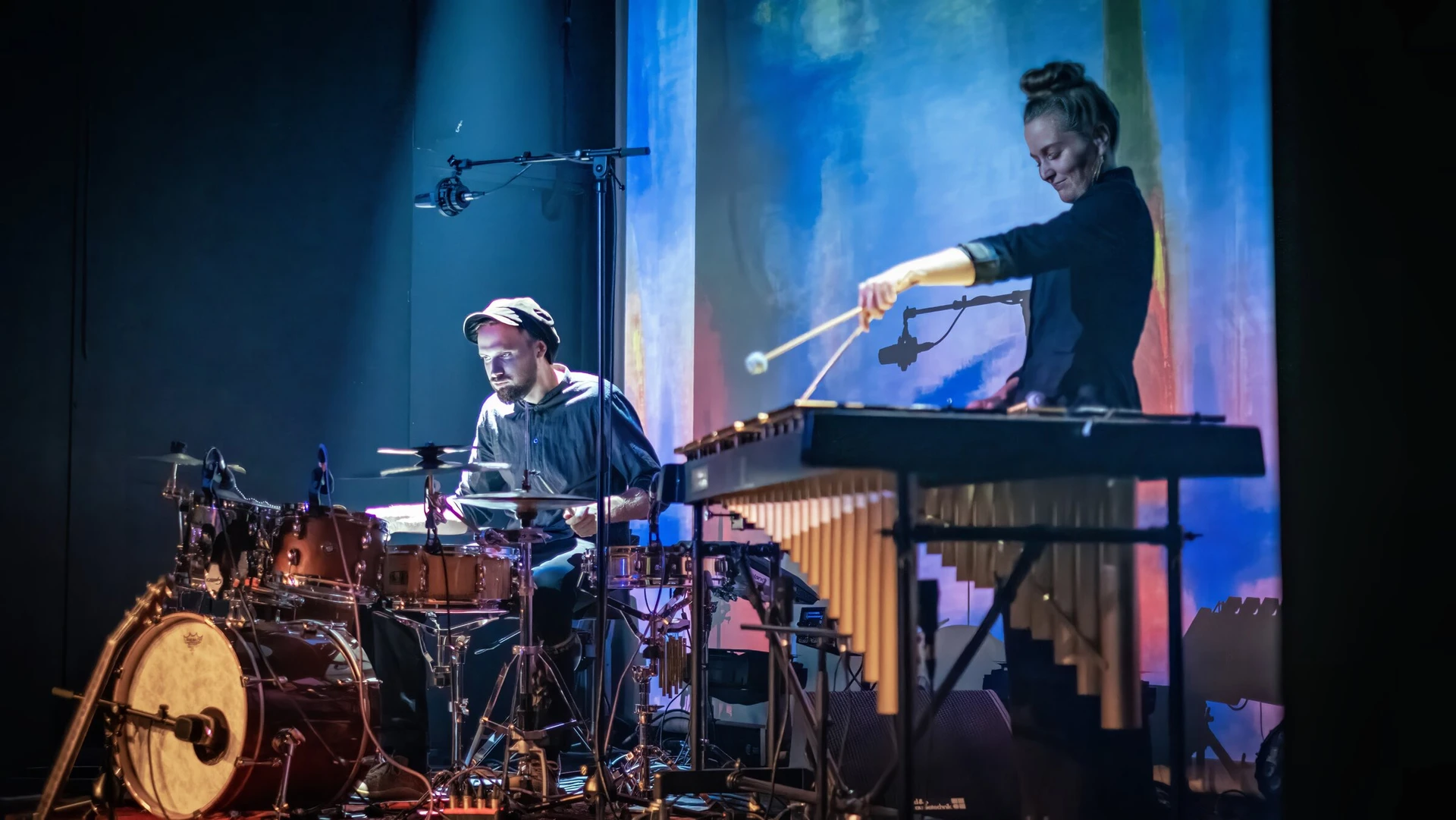 A drummer and a vibraphonist play in front of a screen of soft blue and red painterly light.