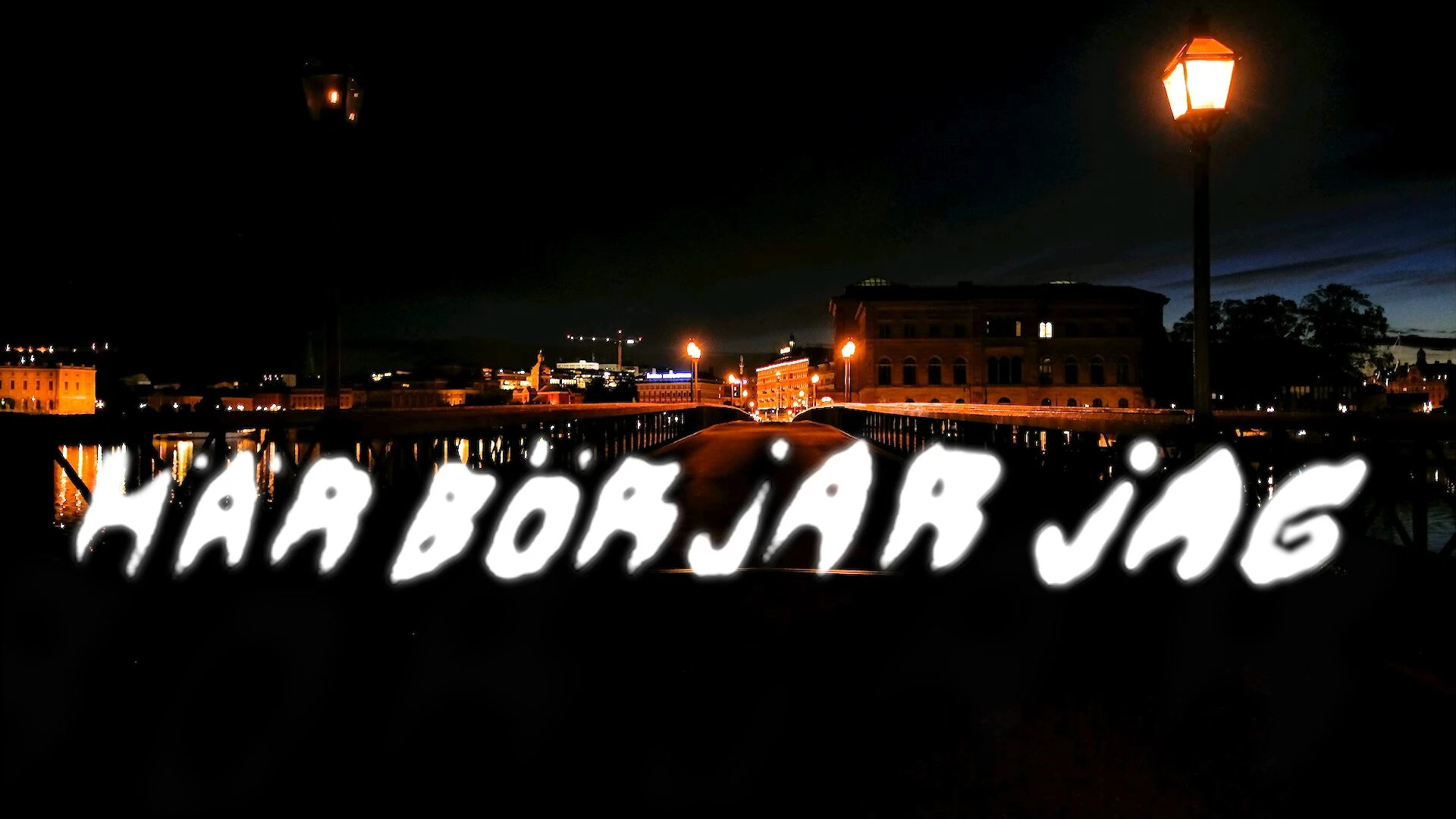 Här börjar jag, written in rough white light letters on a quay at night, the lit waterfront behind.