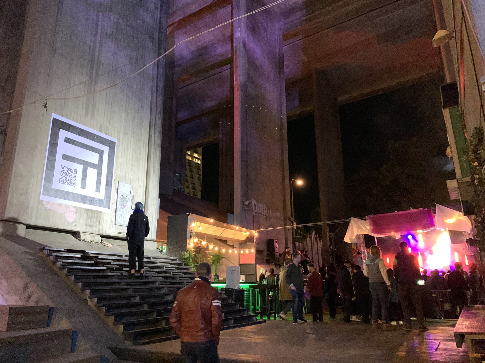 A black and white AR marker several metres across covers a concrete pillar under Skanstullsbron at night, above a dark stairway and the lit market stalls and stage of Trädgården.